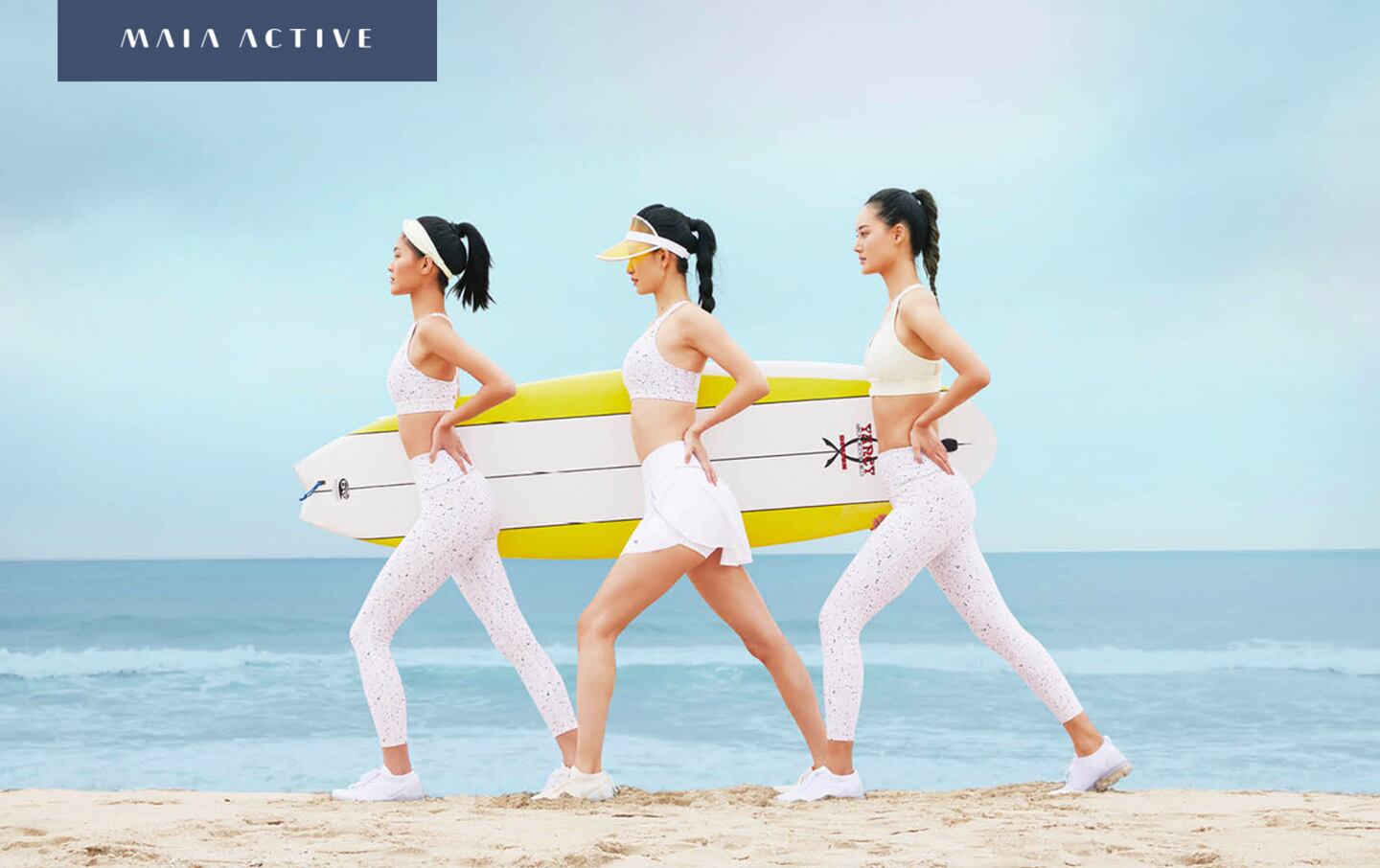 Three models on the beach hold a surfboard while wearing Maia Active clothes.
