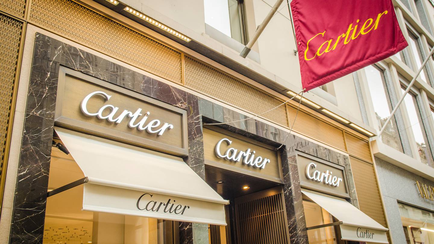 The exterior of a Cartier store in London.  Shop signs are displayed above the door and on a red banner that hangs outside.