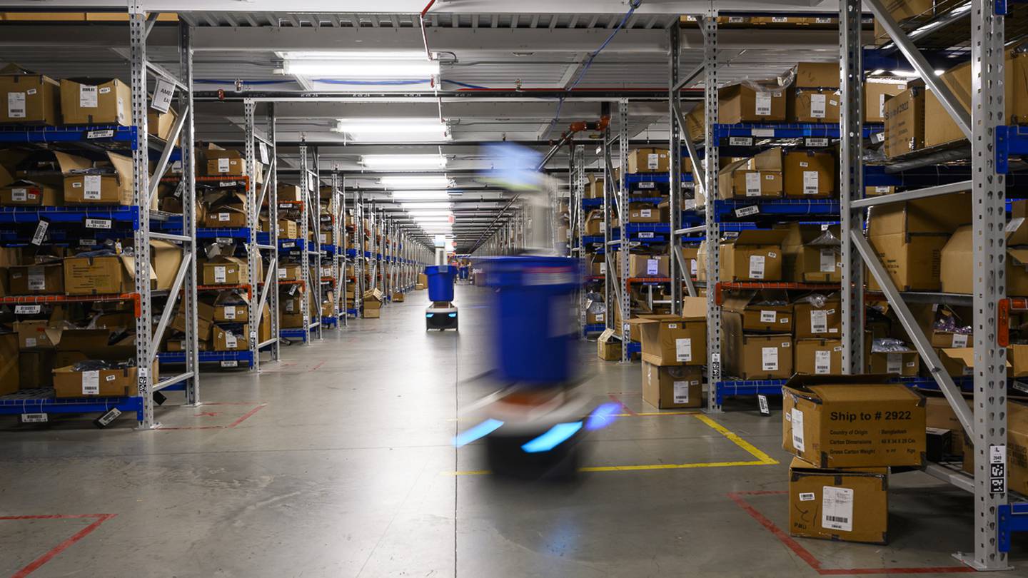 Two small robots are transporting blue bins in a warehouse full of boxes.