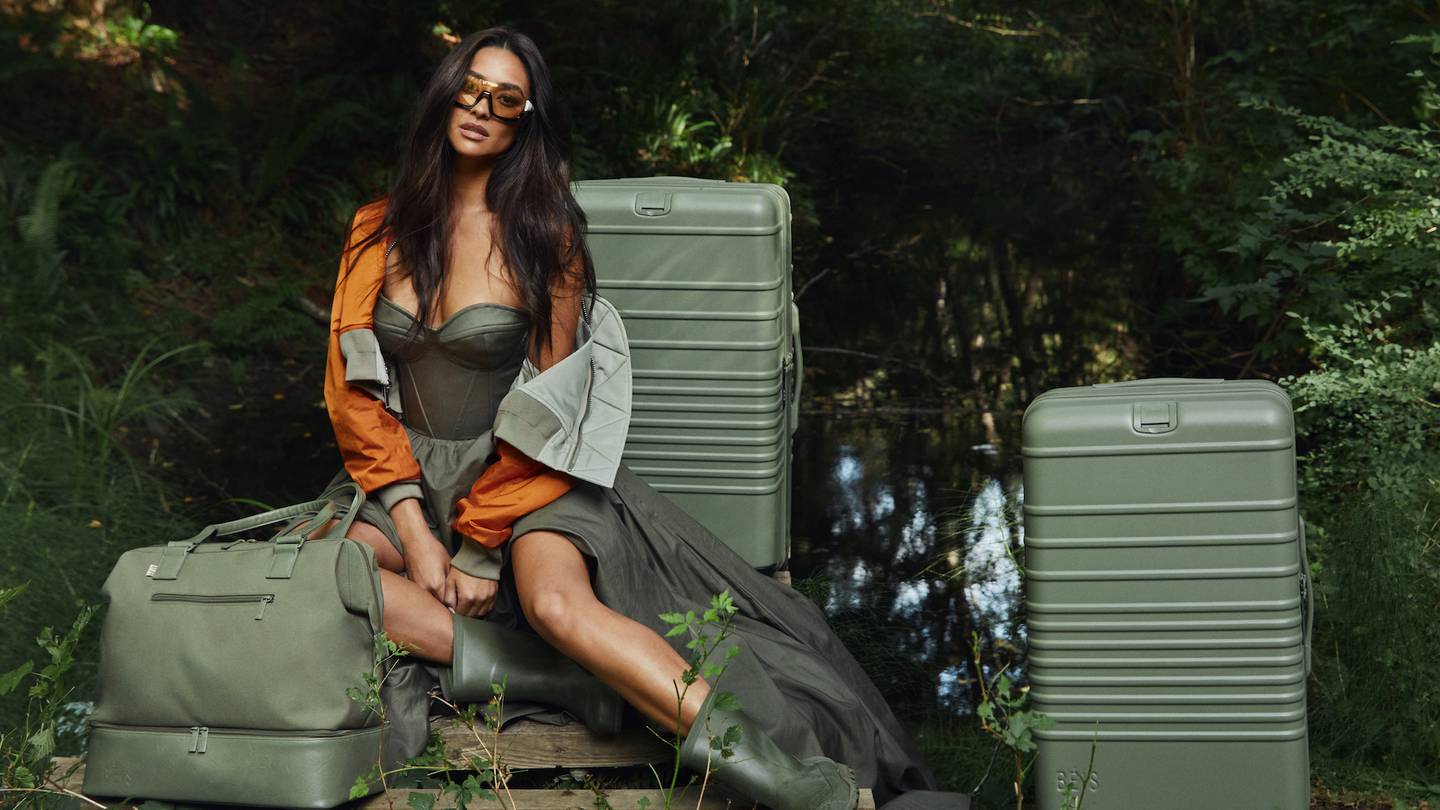 Actress Shay Mitchell sits among two olive-coloured suitcases and weekender bag from her brand Béis.