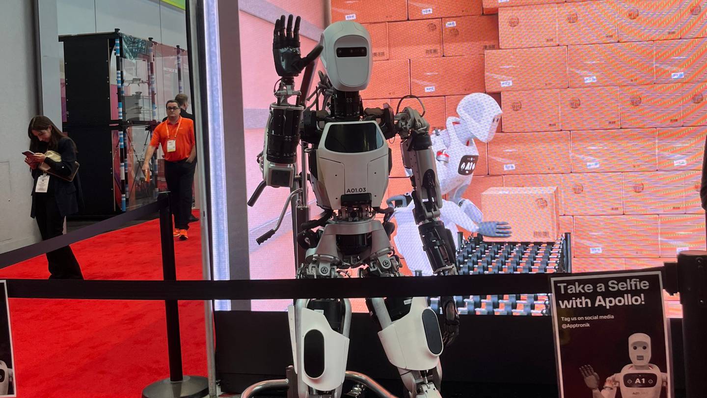 A humanoid robot waves at the camera in front of a screen showing a simulation of a robot stacking boxes in a warehouse.