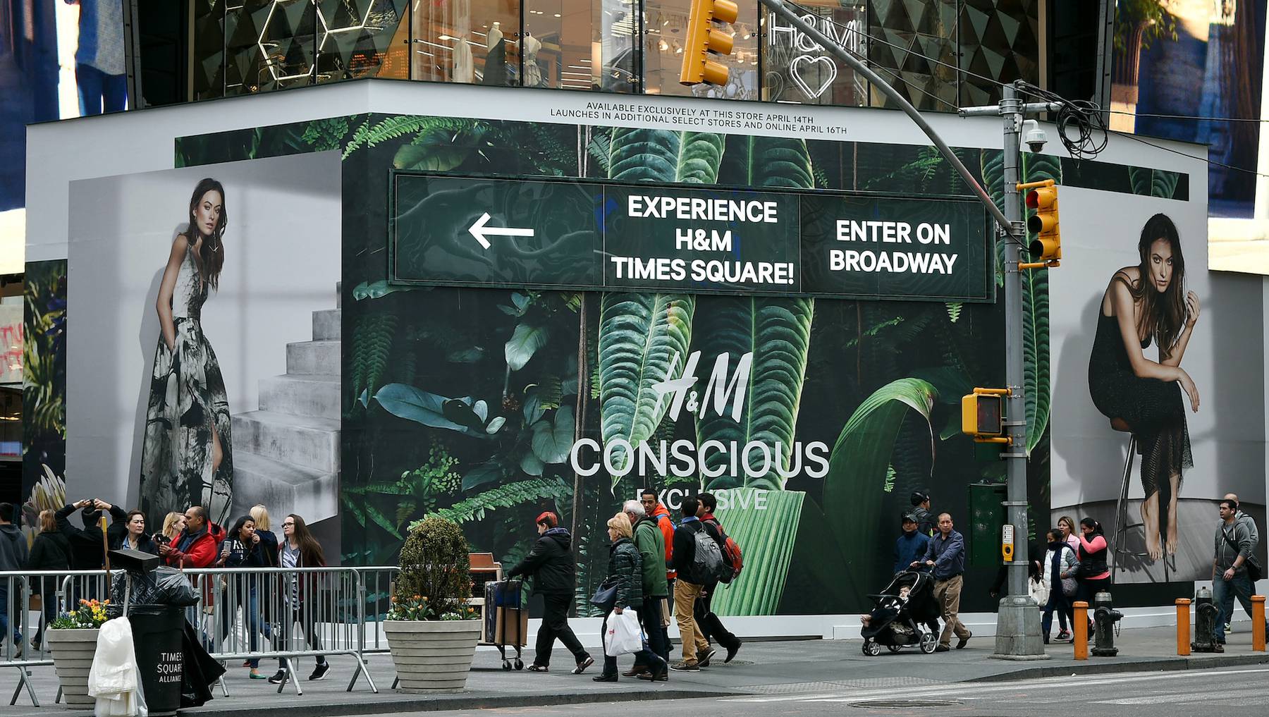 Groups of shoppers pass by on a busy street outside an H&M store in Times Square, New York. The outside of the store is covered by a huge billboard that stretches around the corner. The billboard is covered in green leaves, evocative of a jungle and bears the words "H&M Conscious Exclusive." Either side are two models dressed in clothes from the collection.