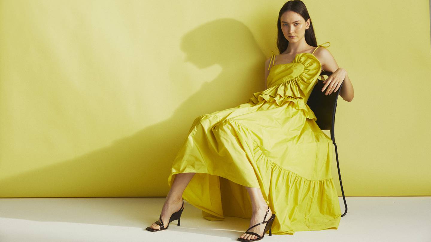 Model in a yellow dress sitting in front of a yellow wall. She wears black strappy sandals.