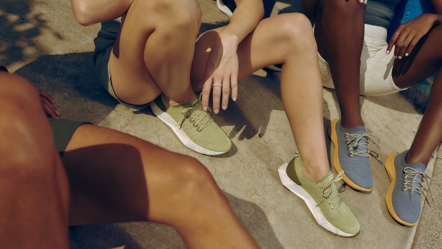 A group of models are shown sitting wearing pairs of green and blue wool Allbirds runners. Only their legs are shown.