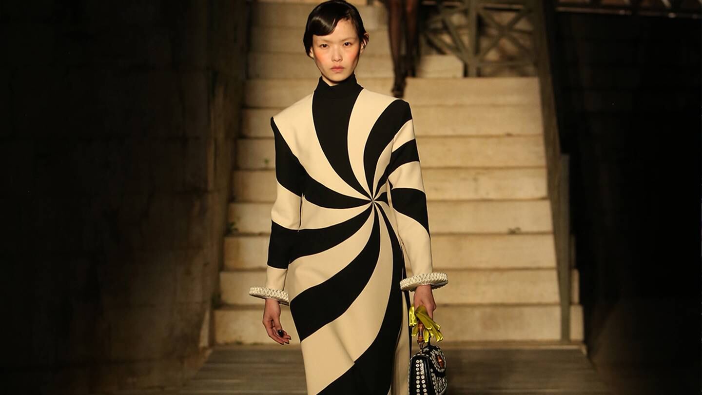 A model walks down the Gucci cruise runway in a black and white swirl dress with a handbag in their left hand.