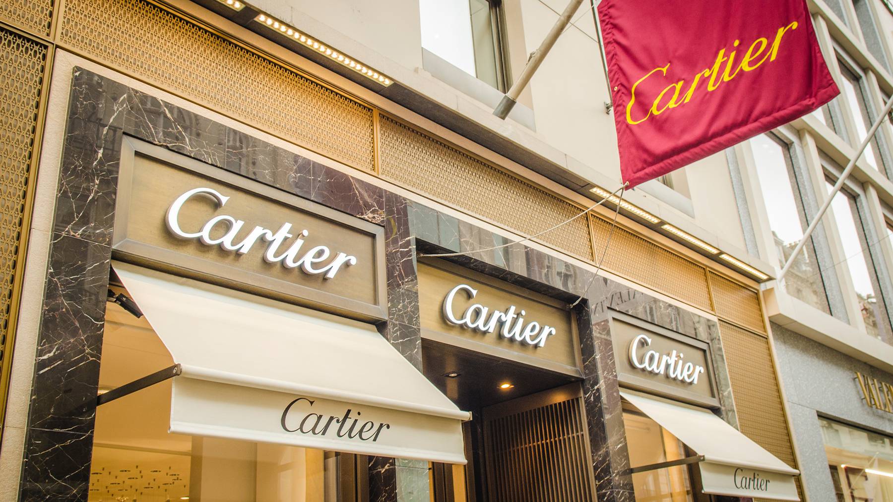 The exterior of a Cartier store in London. Store signs are shown above the door and on a red flag hanging outside.