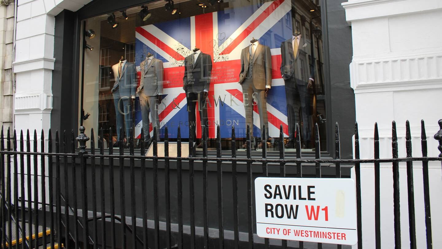 Gieves & Hawkes shop exterior window with suits and a Union Jack flag in the shop front. In front of this is railing with a Savile Row street sign.