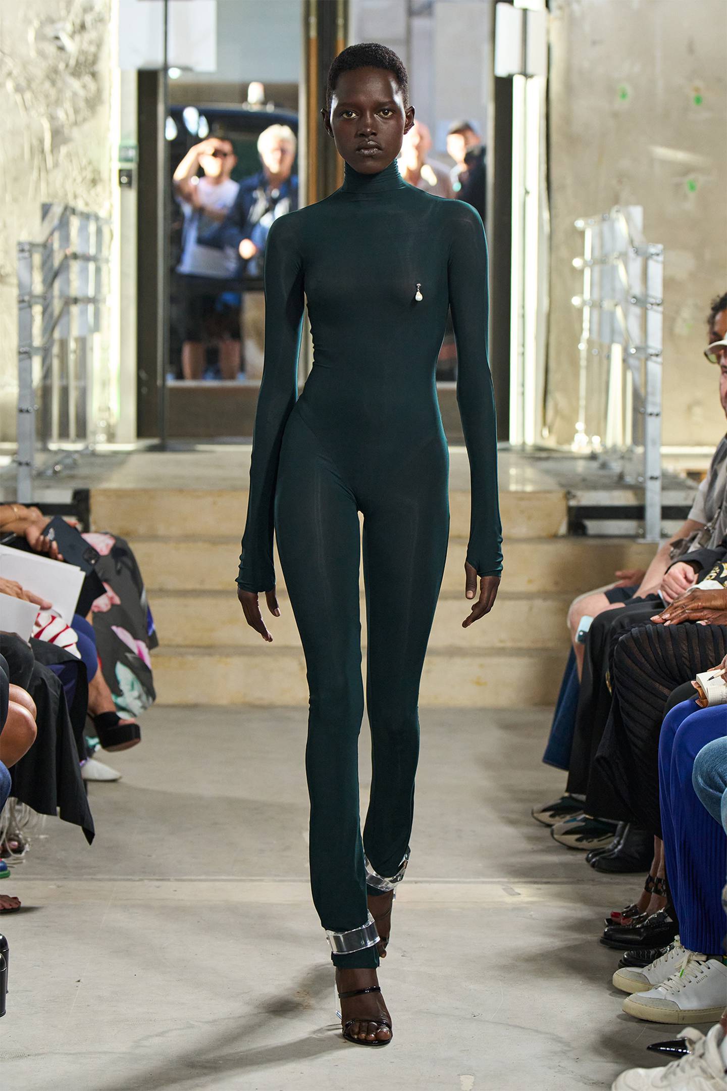 A model wears a dark green emerald catsuit with black heeled shoes.