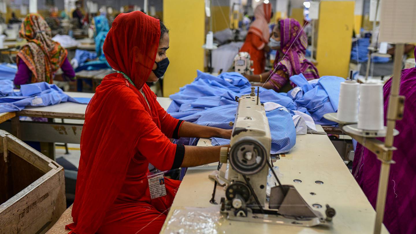 A Bangladeshi garment worker wearing an all red outfit with a black face mask. They sit at a sewing machine using blue materials.