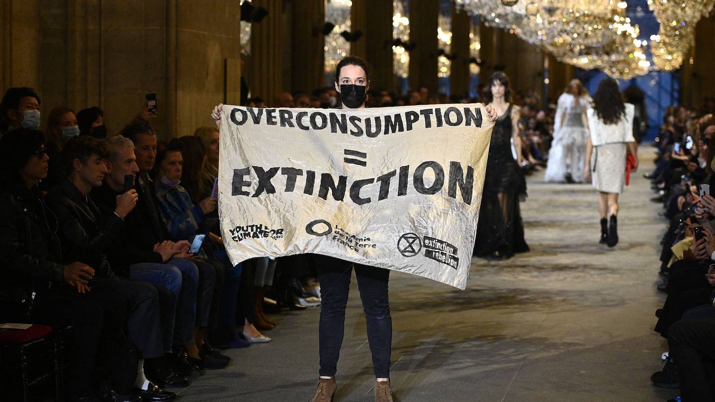 A demonstrator holds a banner saying "Overconsumption = Extinction" as models present creations by Louis Vuitton during the Women's Spring-Summer 2022 Ready-to-Wear collection fashion show as part of Paris Fashion Week at the Louvre in Paris, on October 5, 2021.