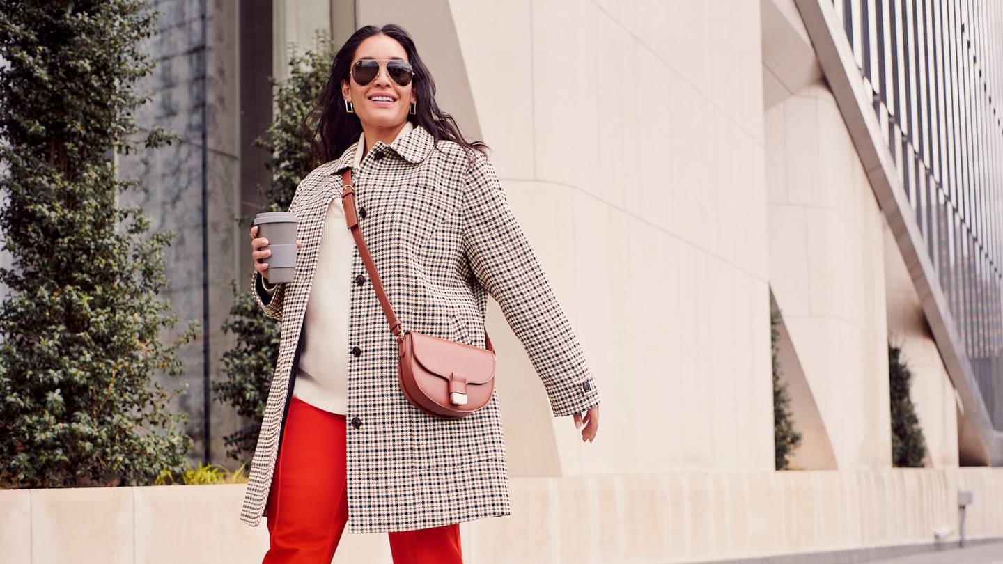 A model in a check coat, off-white sweater and bold red pants walks down a street wearing sunglasses and carrying a cup of coffee.