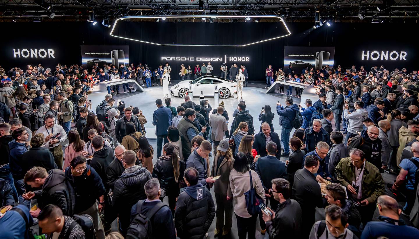 A crowd of people surrounding a central stage where a white Porsche car stands.
