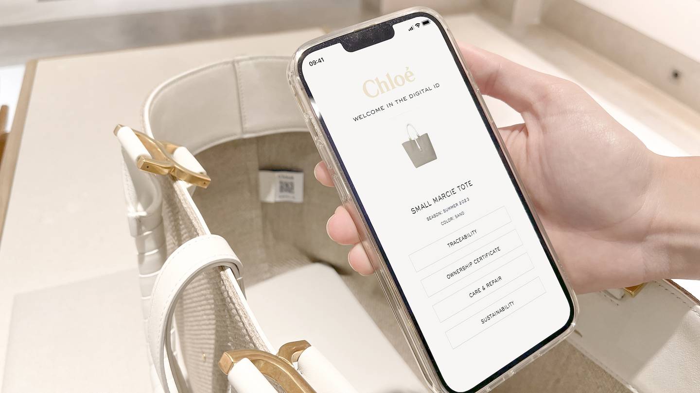 A phone showing a prototype of Chloé's new digital IDs is held above a white handbag.
