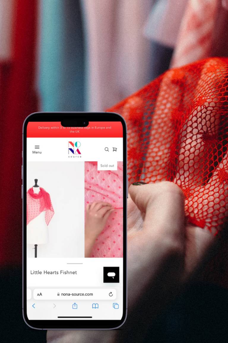 An iPhone with the Nona Source site open is displayed next to a hand touching a red mesh fabric.