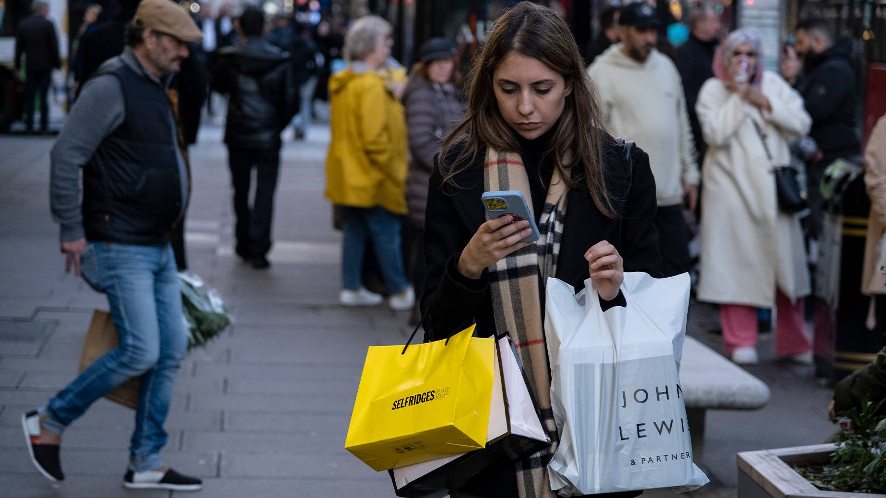 A shopper carrying Selfridges and John Lewis shopping bags, while looking at their mobile phone.