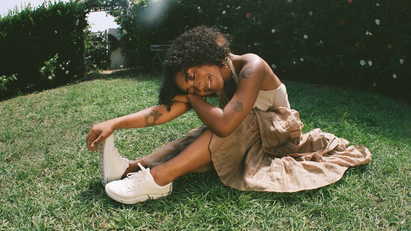A woman sits smiling with her eyes closed on a patch of grass surrounded by bushes. She is wearing a white sleeveless top, brown skirt and white sneakers. She is reaching towards her feet and resting her head on her arms.