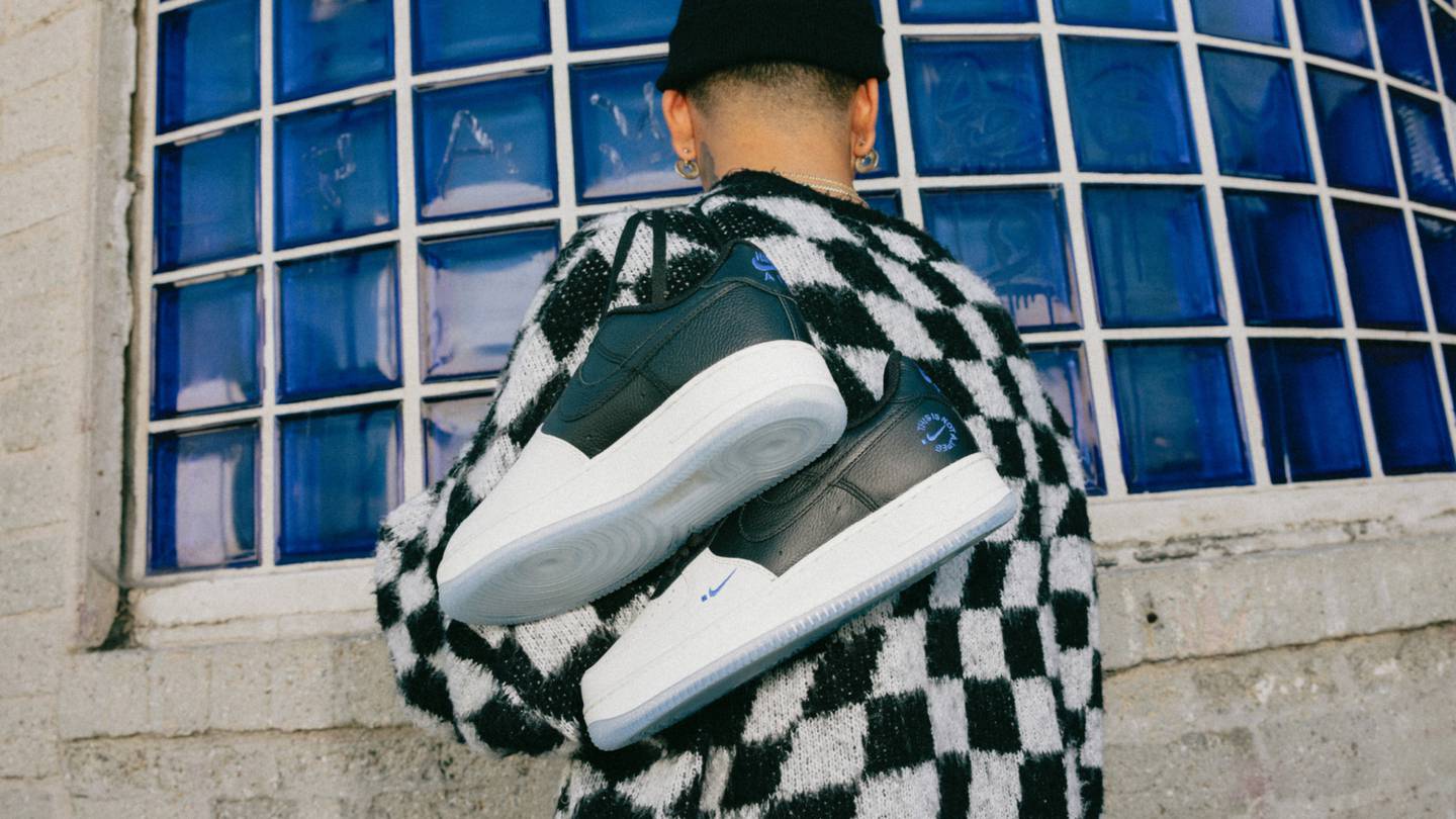 A man in a checked sweater dangles a pair of Nike Air Force 1 TINAJ sneakers over his shoulder.