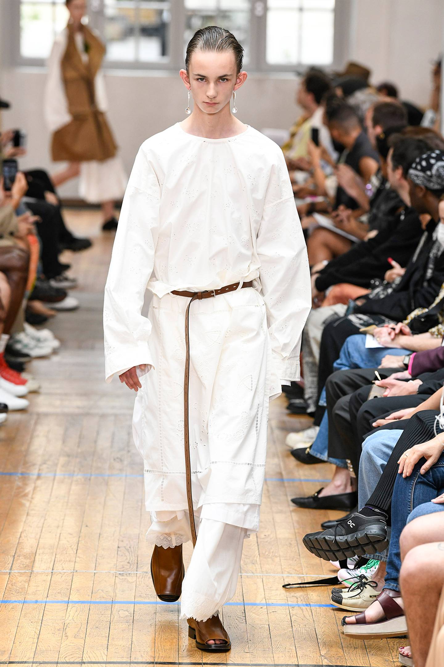 A model wears a relaxed fit white outfit with a long shirt and brown long belt and shoes.