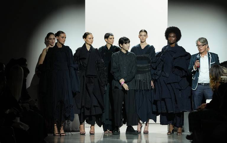 A designer stands with a group of models wearing their black draped designs on a brightly lit catwalk stage.