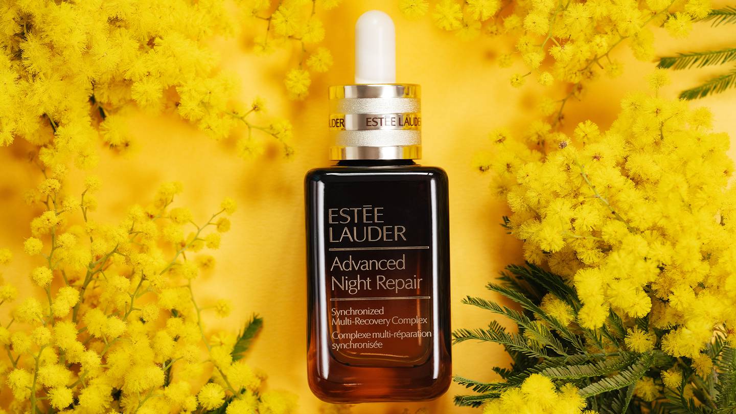 Estée Lauder advanced night repair serum bottle laying on a yellow background with yellow flowers surround it.