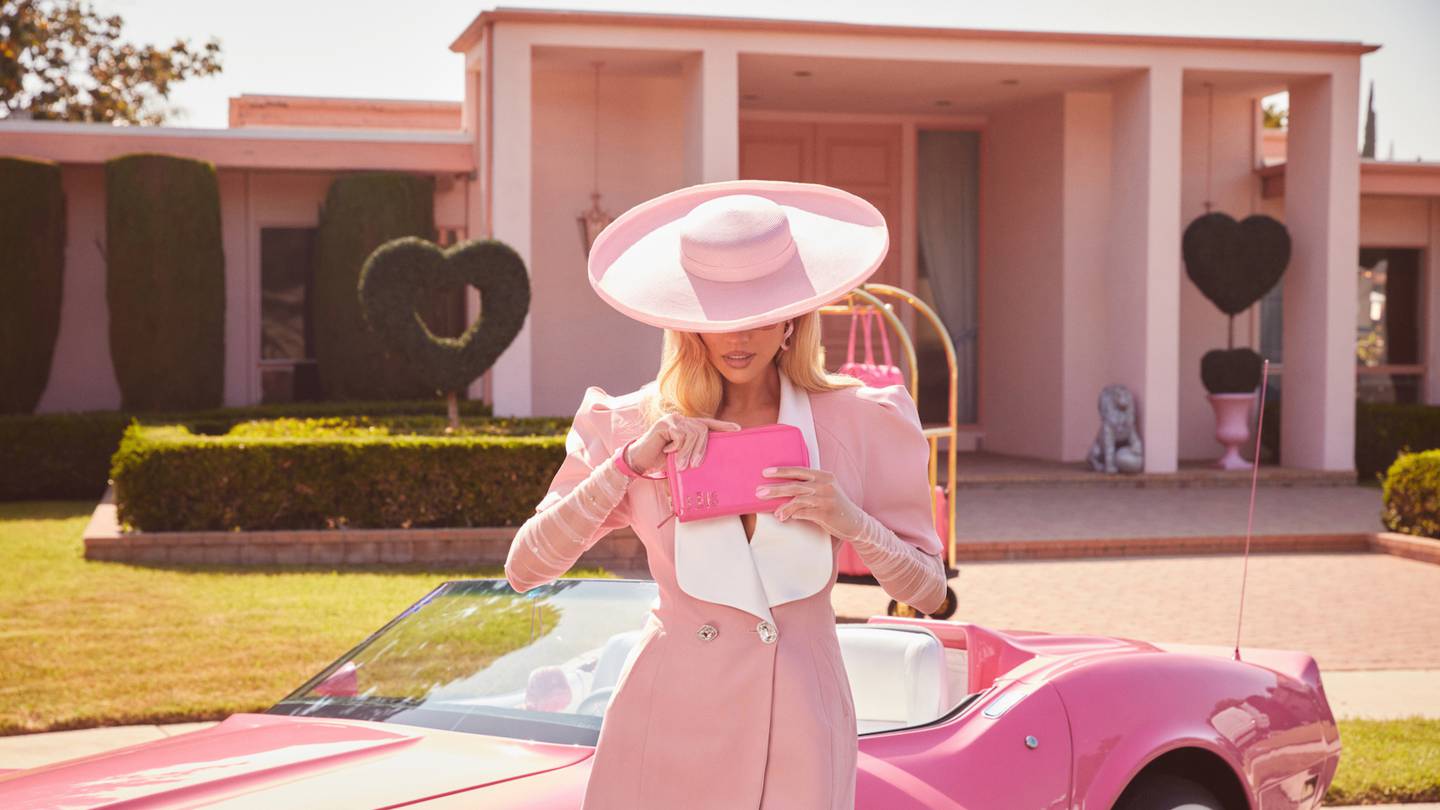 Béis founder Shay Mitchell dressed in a pink suit dress and hat, holds a pink Béis wallet standing in front of a pink convertible car, in a campaign for the Béis and Mattel 'Barbie Pink' colourway collaboration in May 2023.