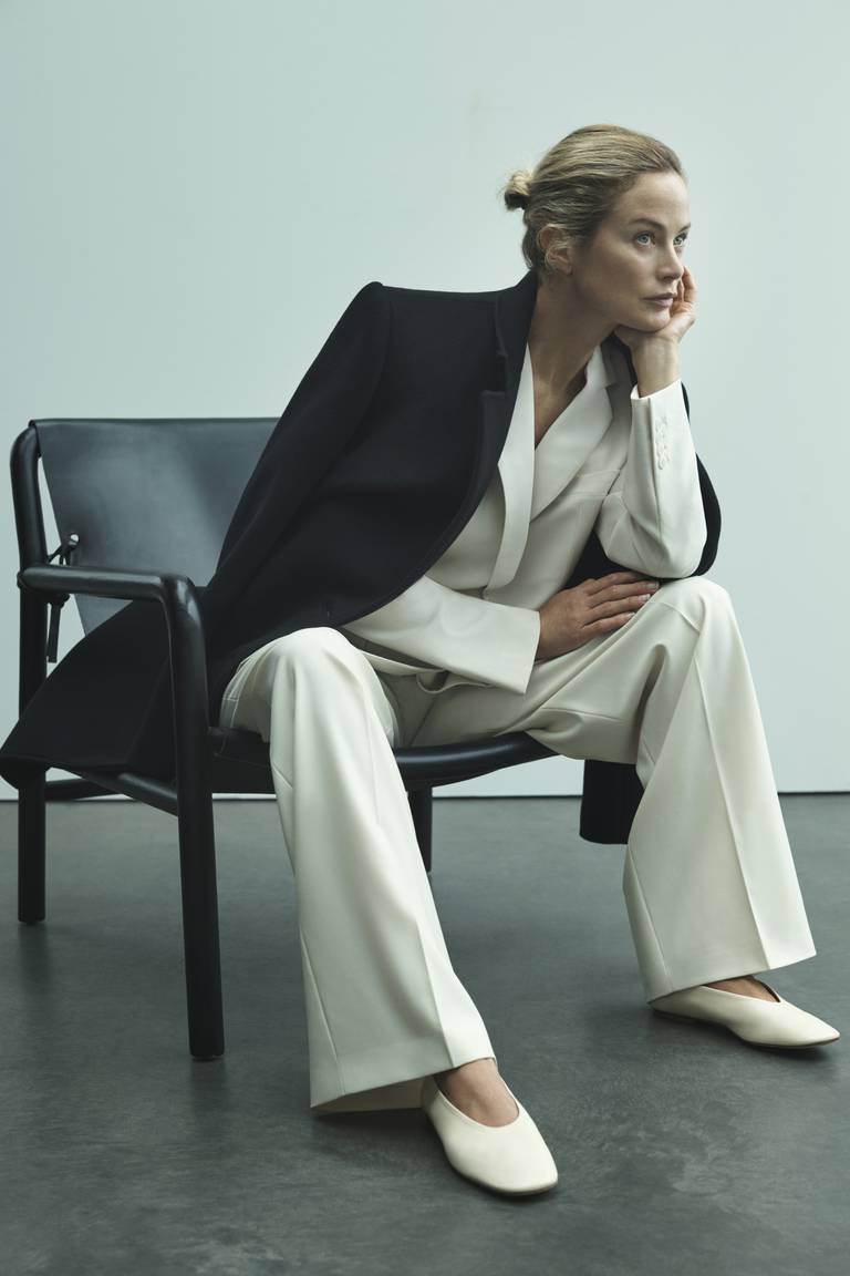 A woman in a white suit is hunched over in a chair with her face propped by her hand.