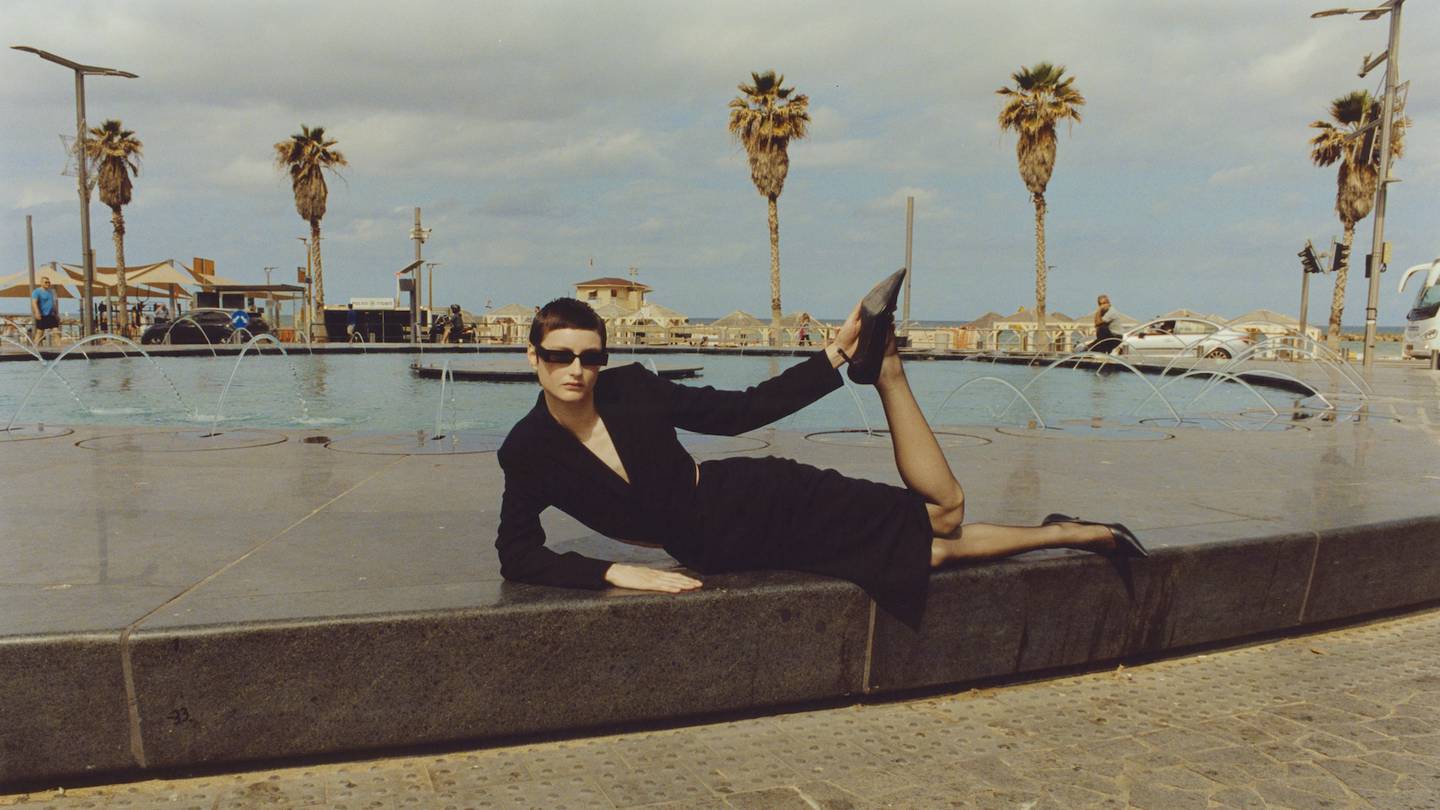 A woman sits by a sunny fountain wearing a sharp tailored black jacket and pencil skirt from the Julie Pellipas brand Better.