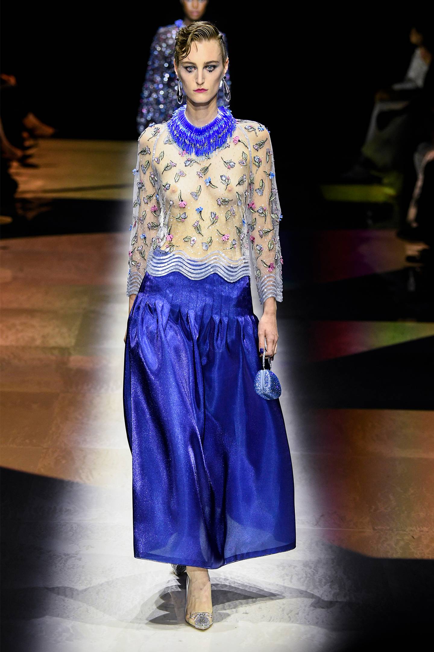A model wears a mesh embellished long sleeved top, with royal blue details, and a satin blue maxi skirt paired with high heeled shoes.