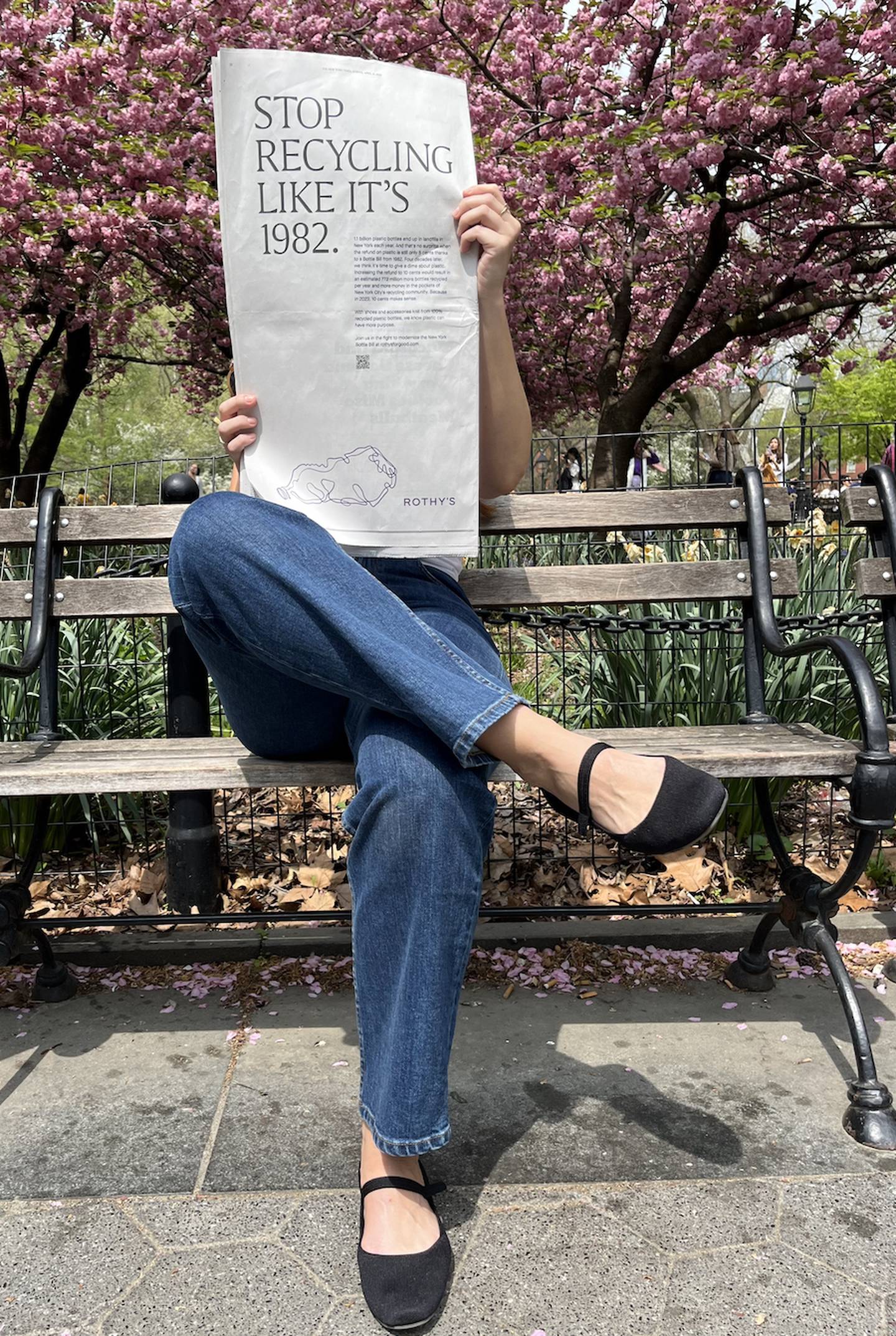 A woman sits under a cherry blossom tree wearing Rothy's shoes and reading a paper with an ad for the New York Bottle Bill.