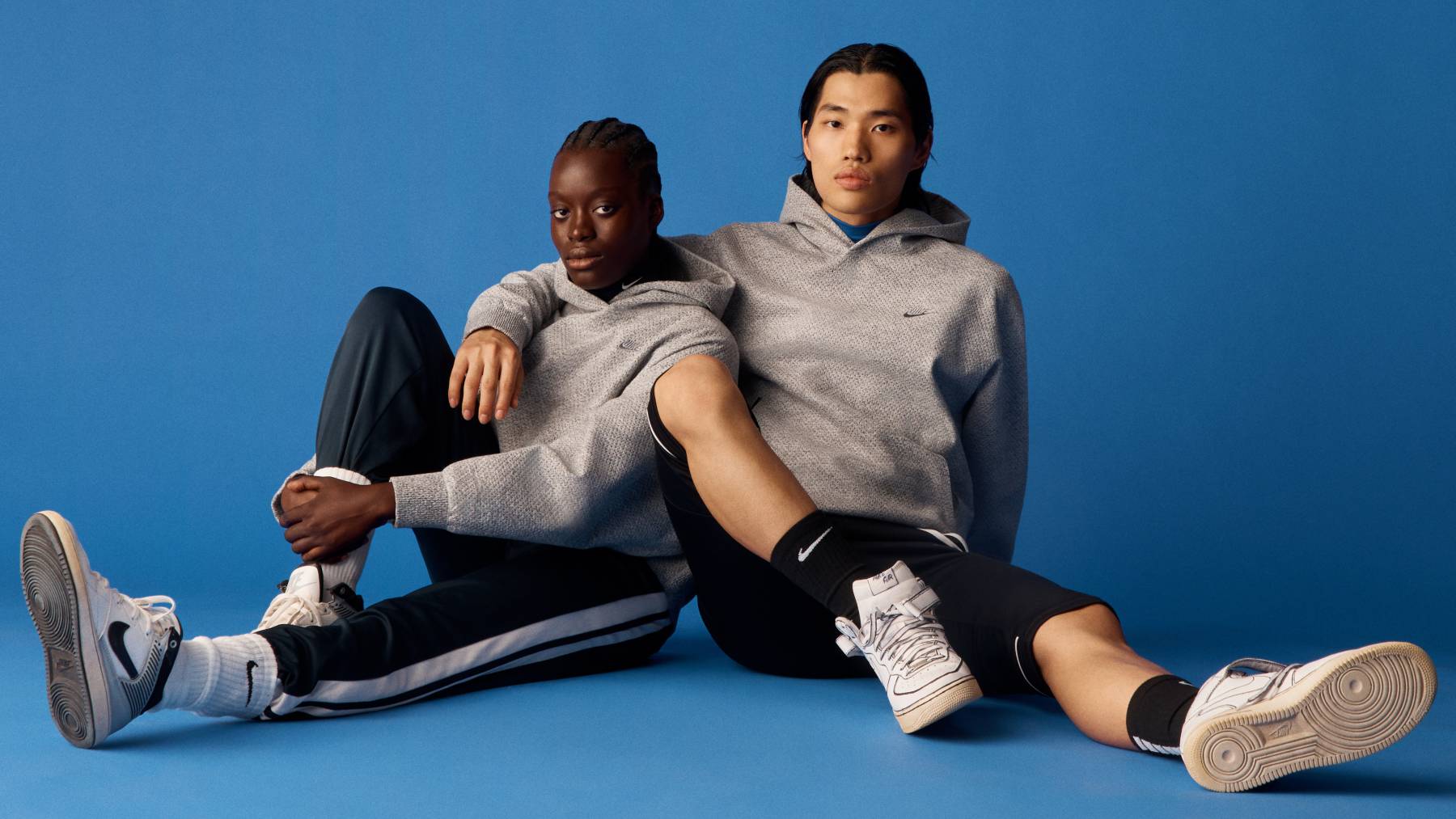 Two models wear grey Nike Forward hoodies against a blue backdrop.