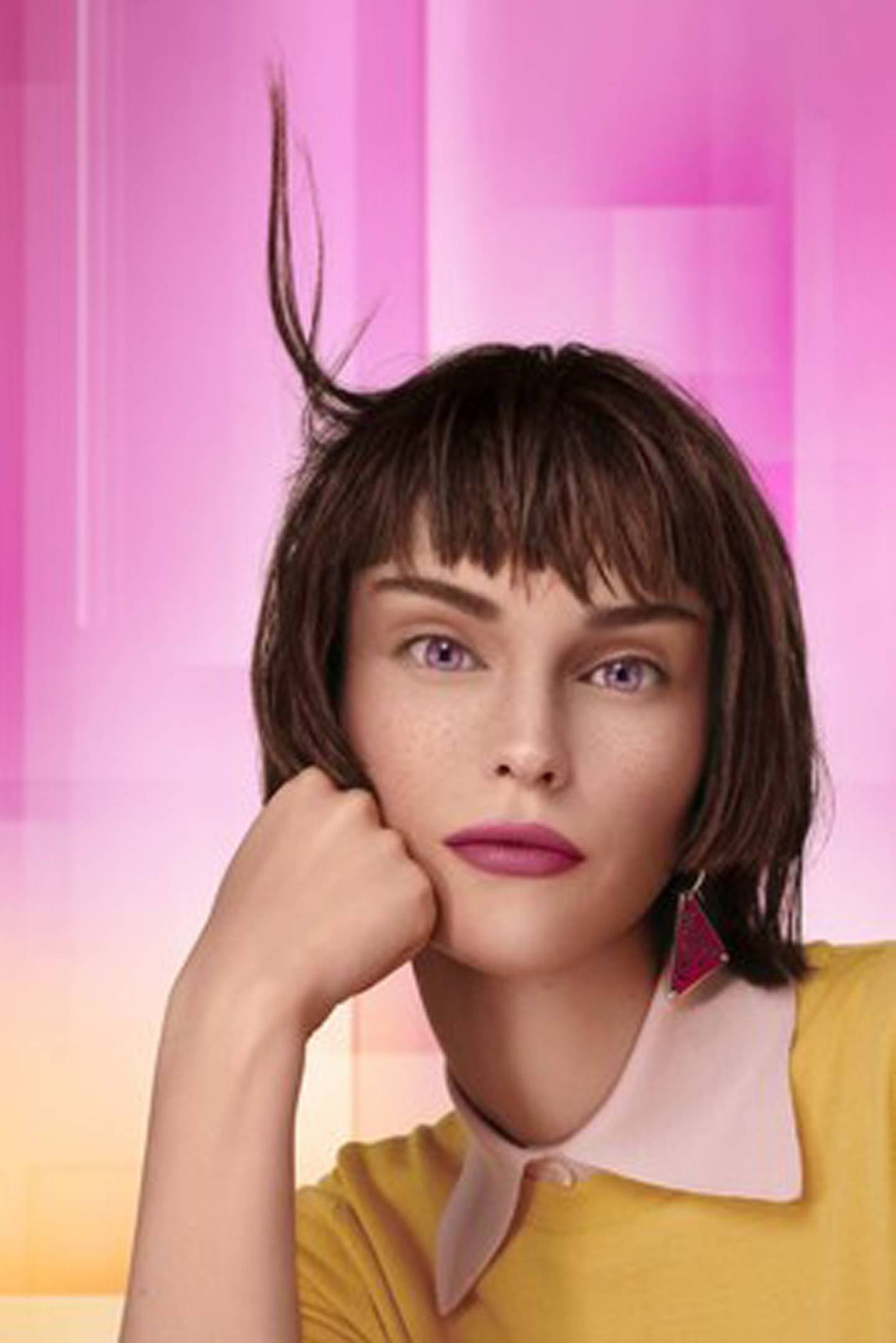 A virtual model with a shaggy bob haircut and bangs leans her head on her hand and looks straight at the viewer.