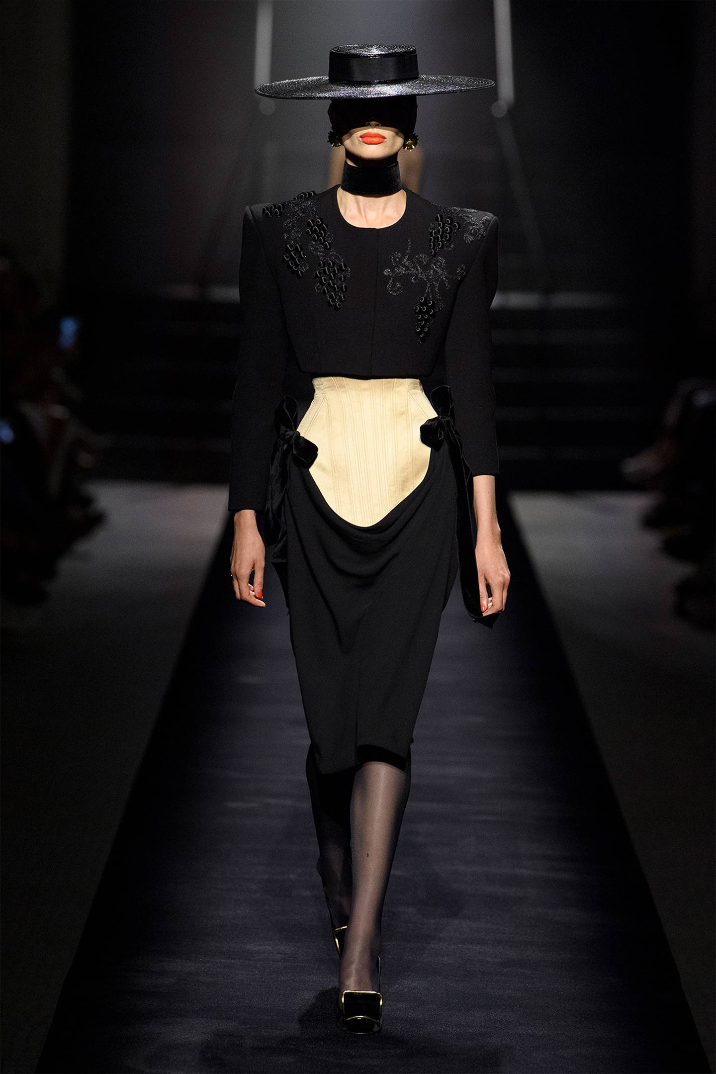 A model wears a black hat and black long sleeved dress with black tights and shoes and a gold belt.