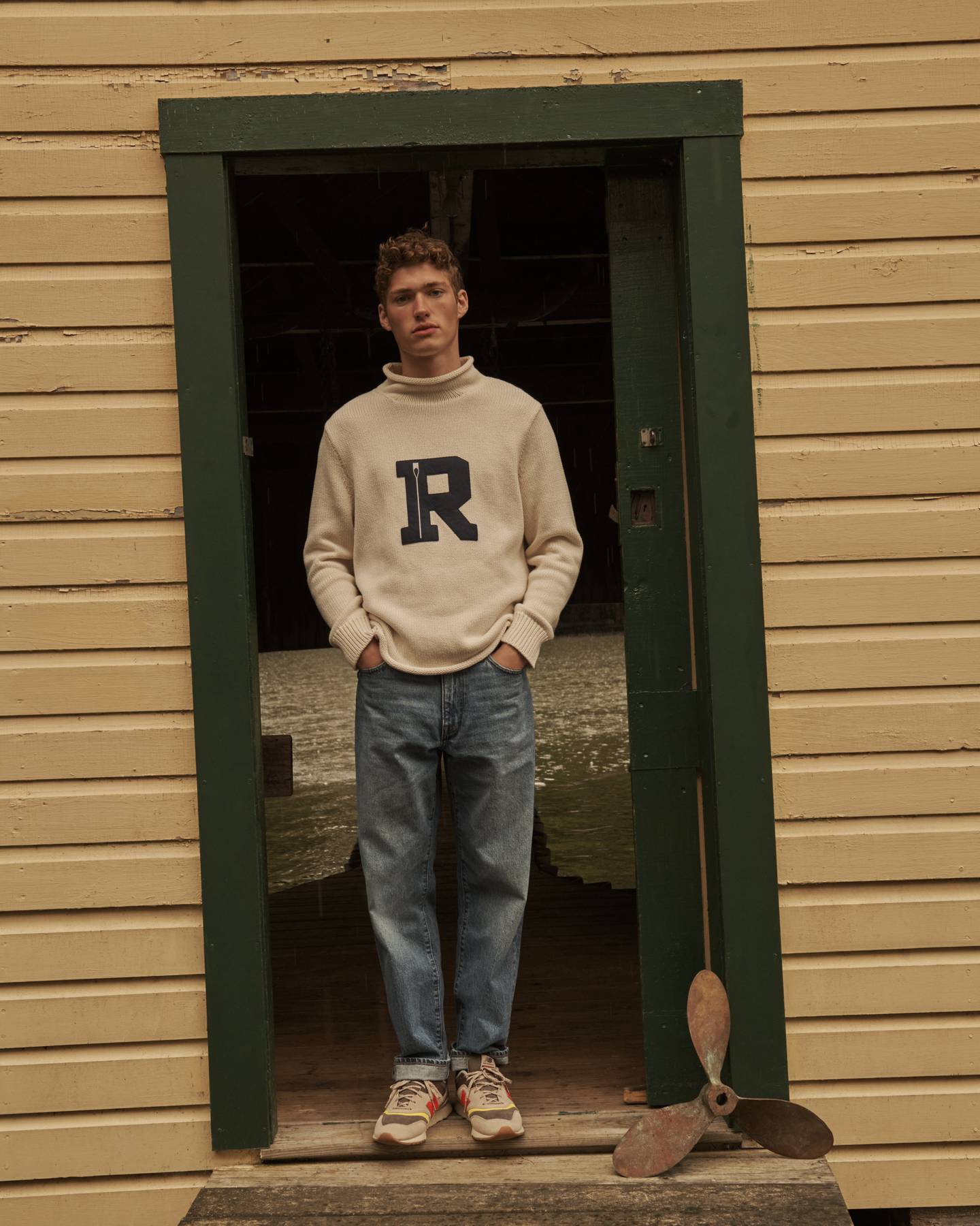Man stands in doorway wearing mockneck sweatshirt with the letter 'R'