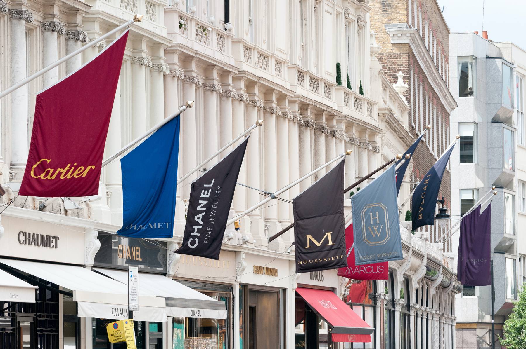 The exterior of luxury stores on Bond Street in London. Flags showing brands including Cartier and Chanel are above the store entrances.