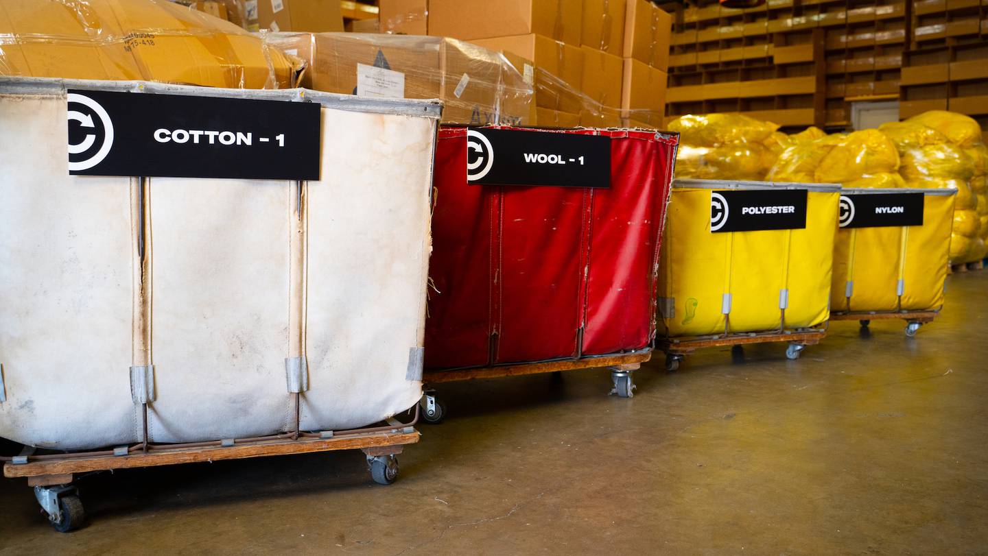 Industrial baskets are lined up in a warehouse. A white one is filled with packages of clothes made from cotton, a red one contains wool and two yellow ones polyester and nylon.