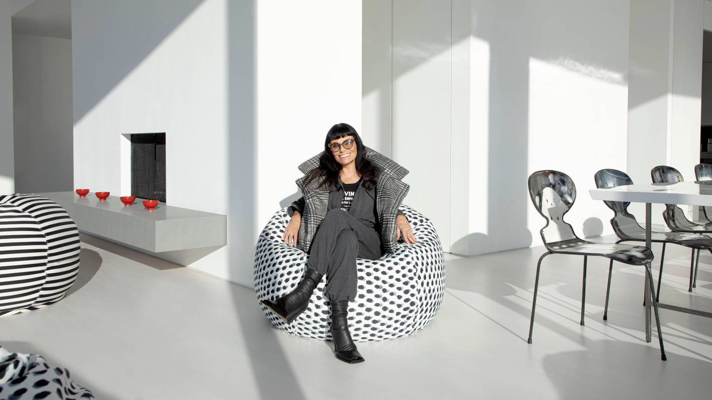 Designer Norma Kamali sits smiling in a polka-dotted chair in a brightly lit room.