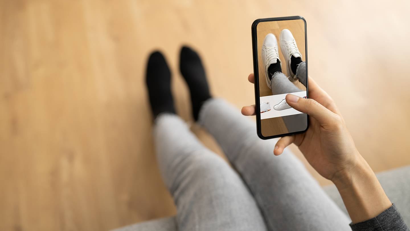 A person uses their mobile phone to use virtual fitting room software to try on white lace up shoes.