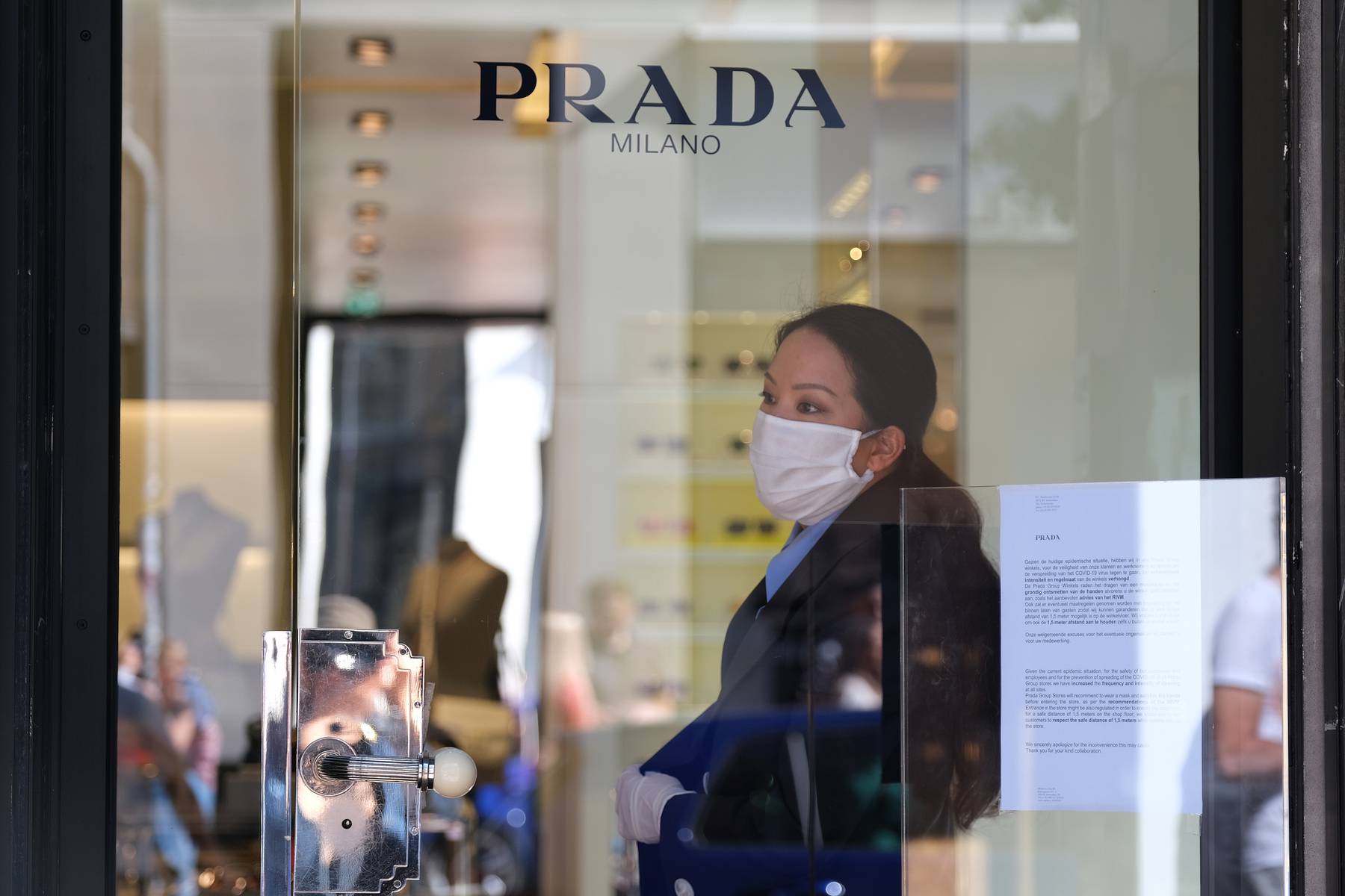 A staff member of Prada, wearing a mask and protective plastic gloves, stands behind a glass door with an advisory on how to shop amid coronavirus