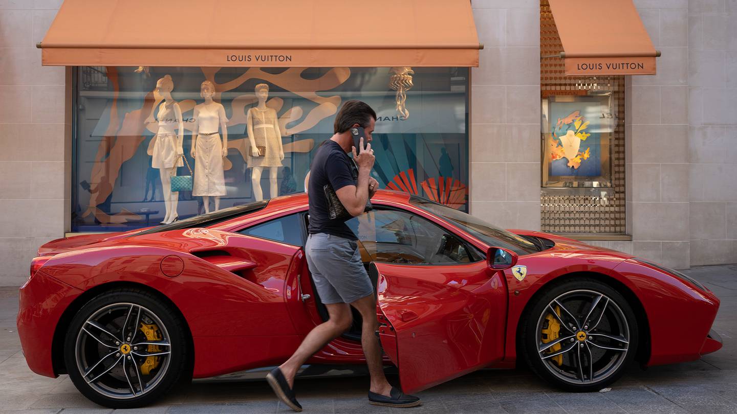 A man on the phone opens a red Ferrari car door. The car is parked in front of the show winds of a Louis Vuitton boutique.