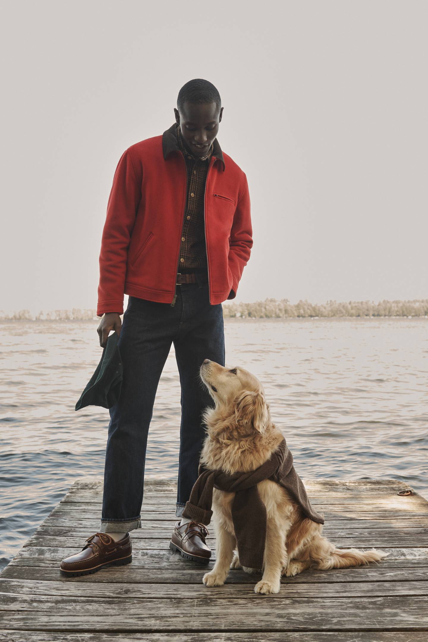 Man in red jacket stands on a dock, looking down to a golden retriever with a sweater tied around its body.