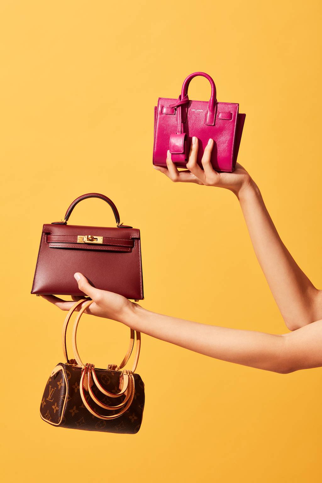 Two hands holding handbags by Louis Vuitton, Hermes and Saint Laurent.