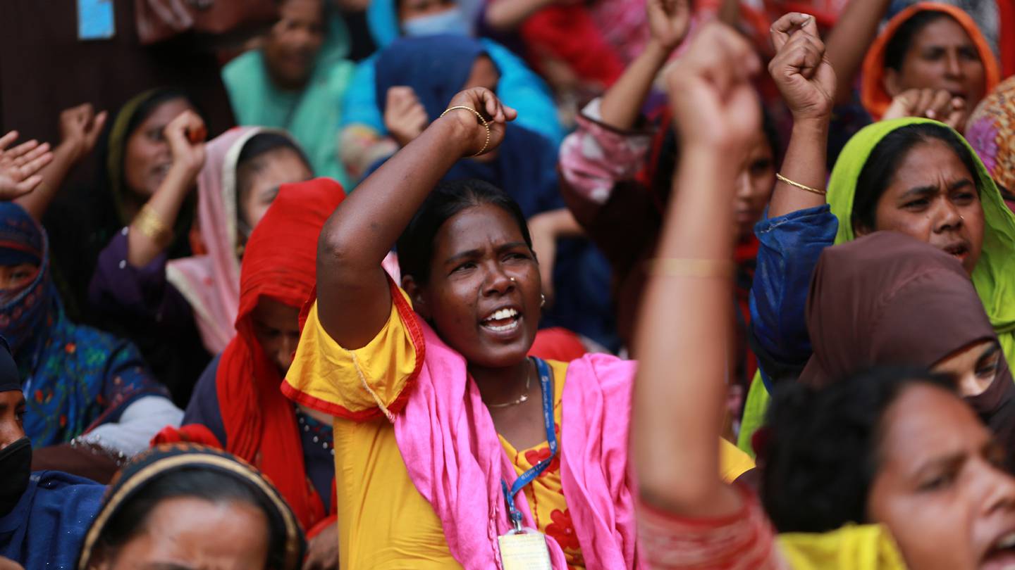 A woman in a bright yellow dress stands in a crowd with her fist raised, part of a demonstration of garment workers in Dhaka, Bangladesh, demanding the payment of due salaries during the pandemic.