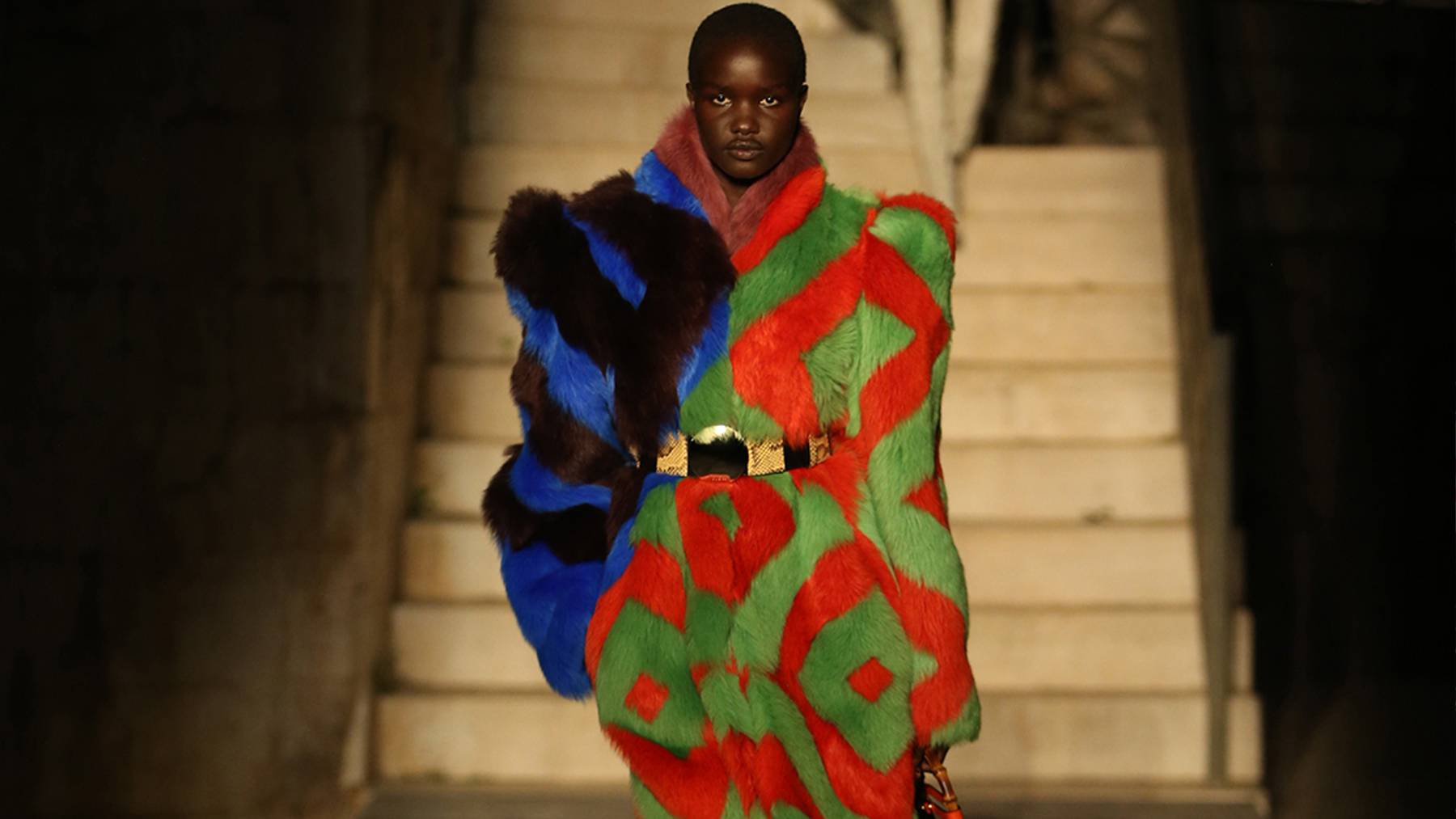 A model walks down the Gucci cruise runway in multicoloured fur belted coat with a handbag in their left hand.