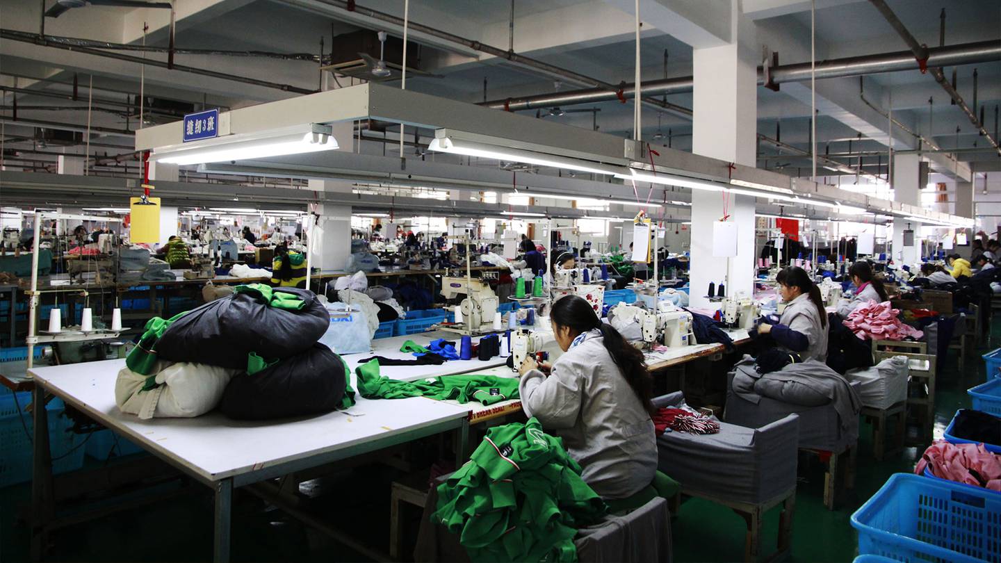 A garment sewing machine floor at a factory in China. Garment workers are stationed at individual sewing machines across large tables with green materials.