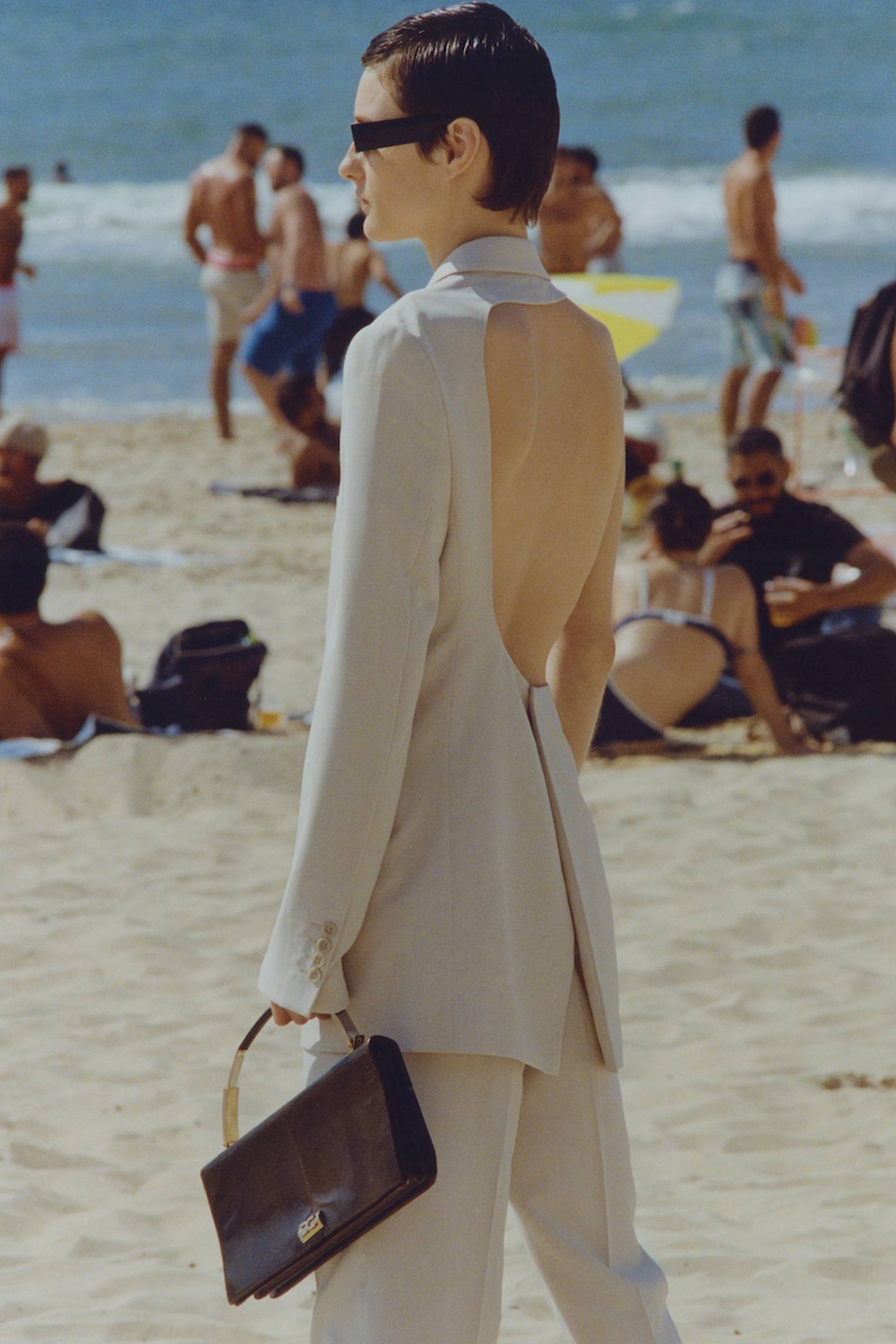 A model in a sand-coloured backless suit stands on the beach.