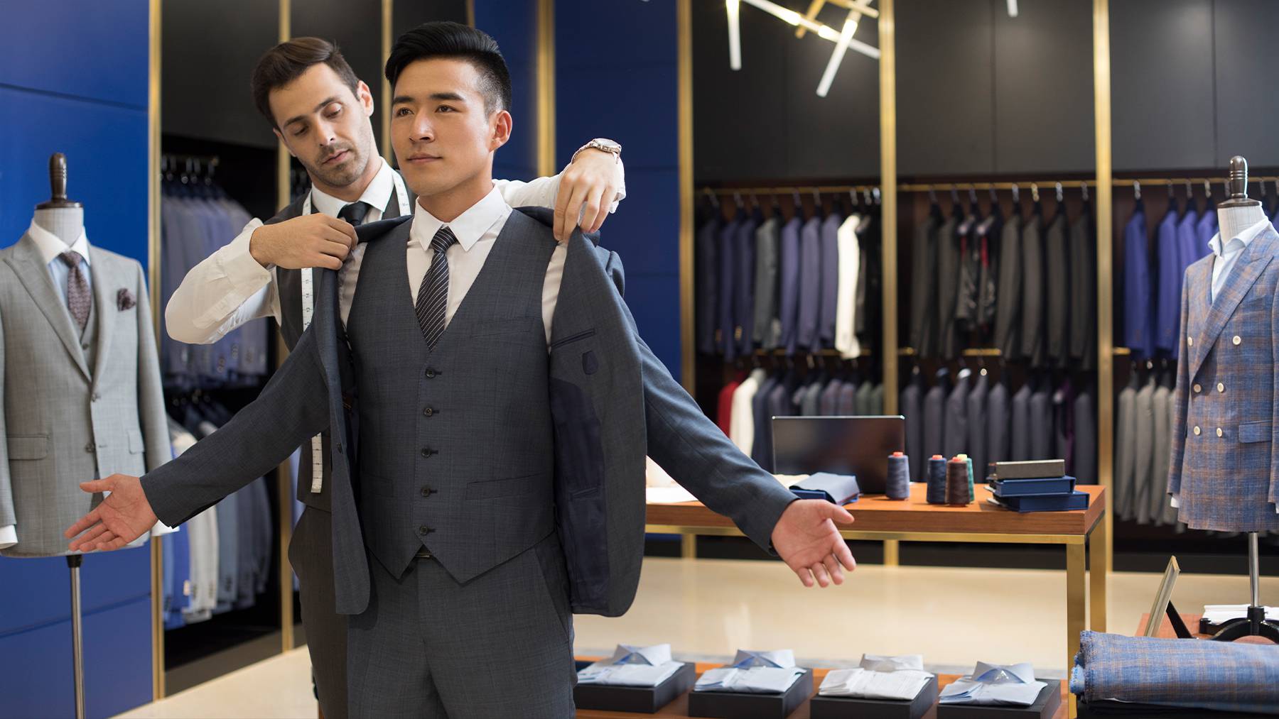 A tailor and customer stand in a tailor boutique. The customer is trying on a three piece grey suit with a white shirt and tie.