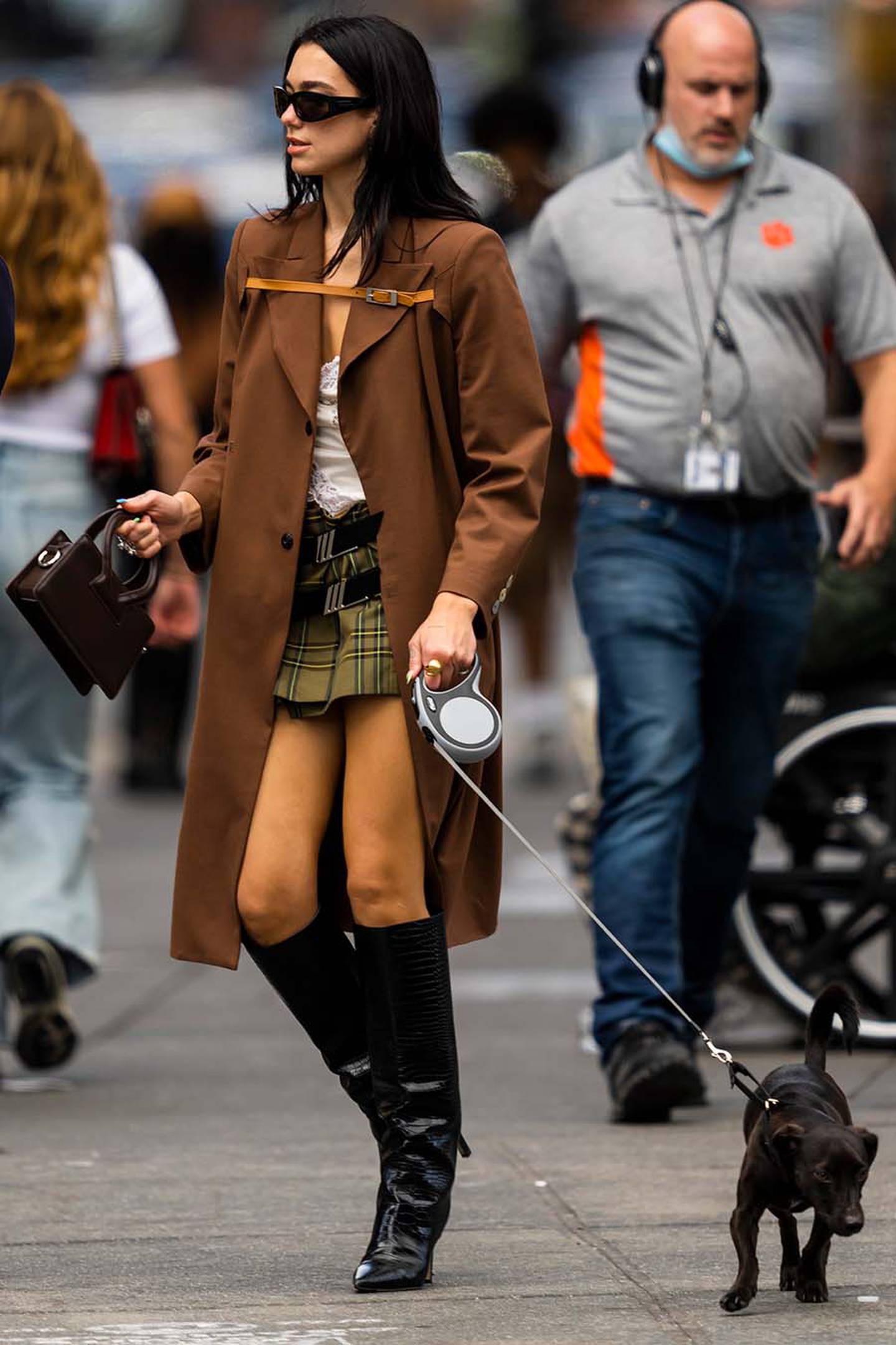 Dua Lipa walking in the street with a small dog. Wearing a long brown coat and Luar brown handbag.