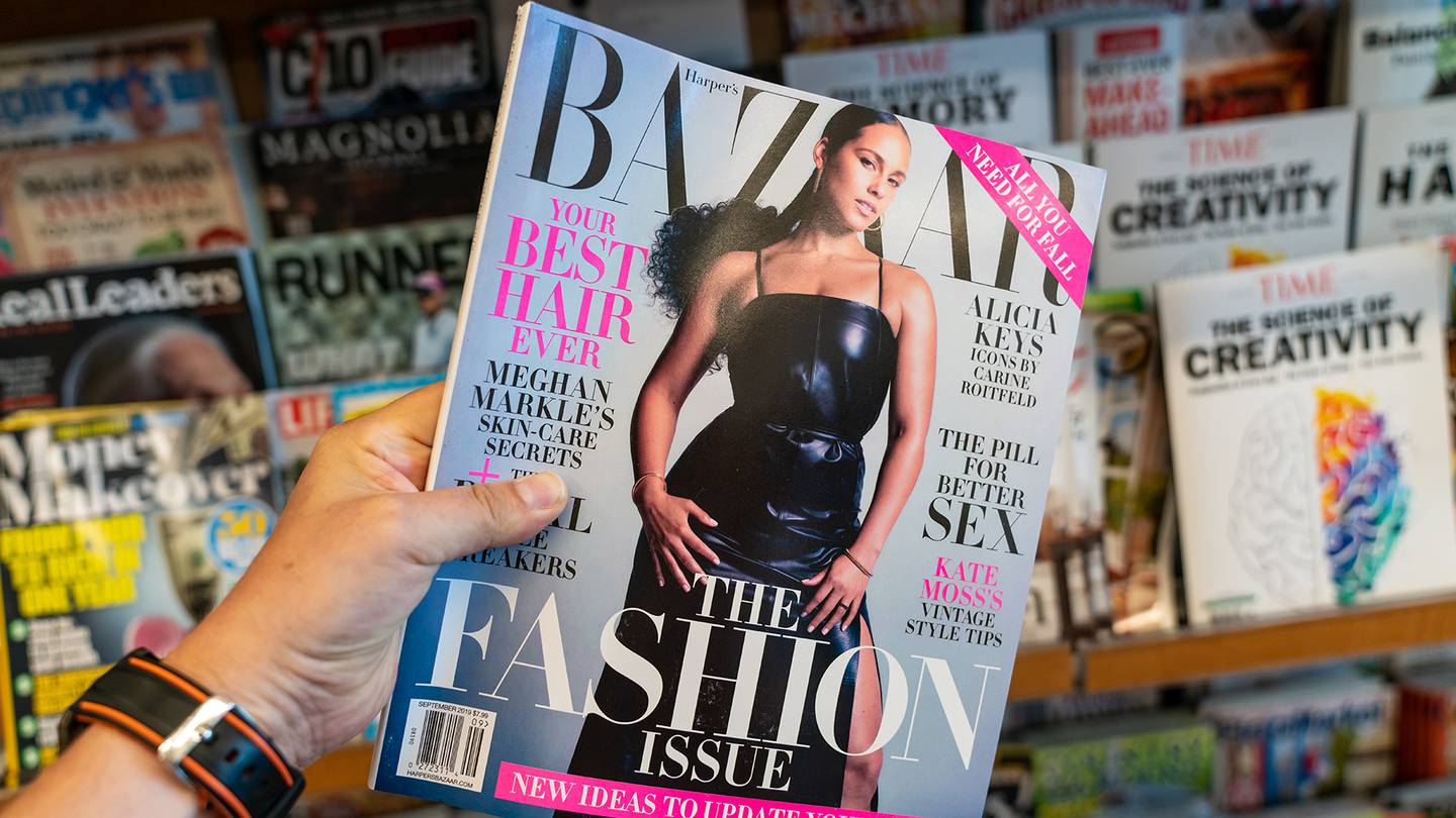 A person holding an edition of Harper's Bazaar magazine at a news stand.