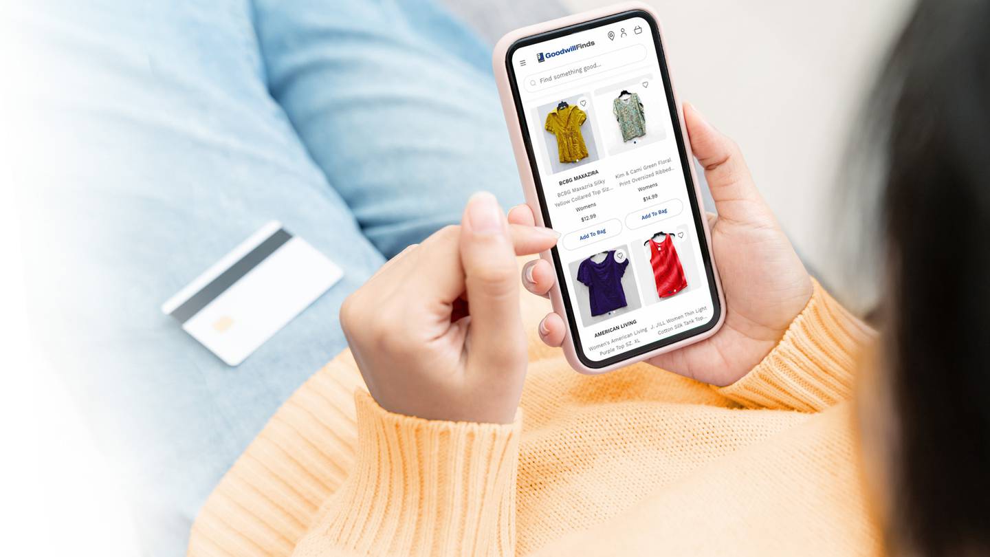 A woman wearing an orange jumper and blue jeans using the GoodwillFinds platform on her phone.