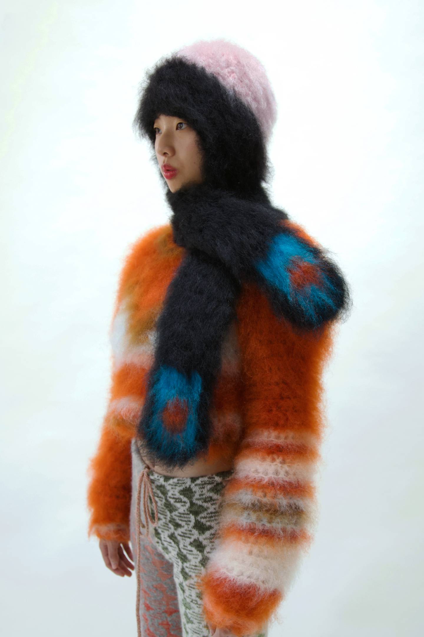 Women wearing a fuzzy hat, scarf and jumper. The hat is pink and black, the scarf is black with bright blue ends and the jumper is bright orange and white stripes.
